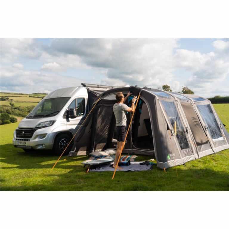 A person setting up an awning next to a camper van on a grassy field with hills in the background.