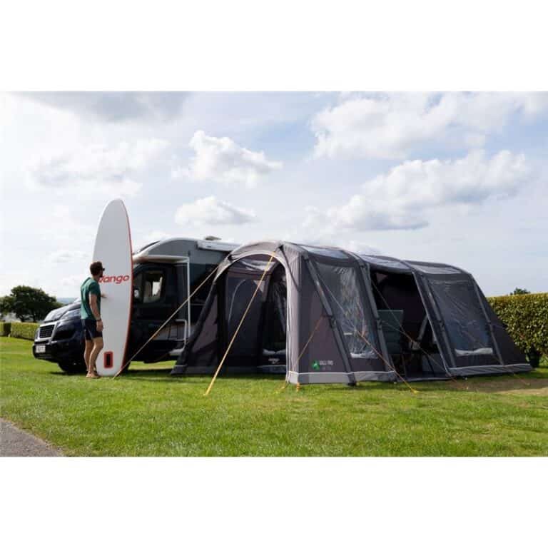 A man stands next to a caravan with a surfboard, connected to a large tent on a grassy campsite.