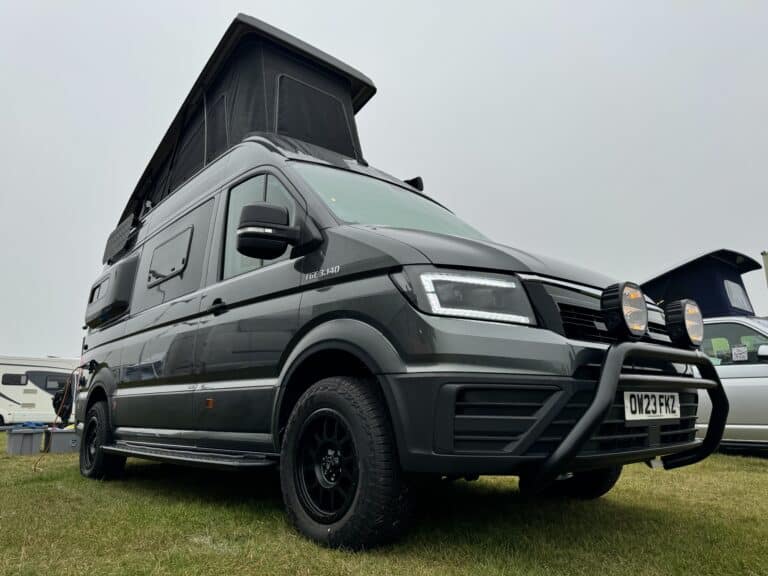 A modern camper van with a pop-top roof parked on grass at an outdoor event.